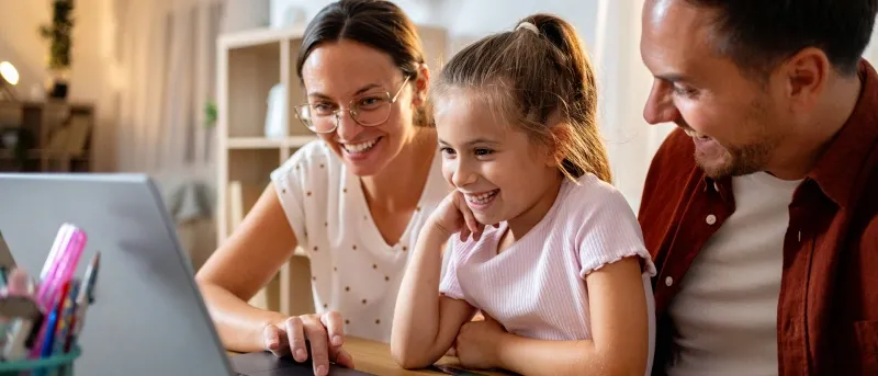 A smiling family, consisting of a mother, father, and young daughter, sits together at a table looking at a laptop screen, with a credit card visible on the table.